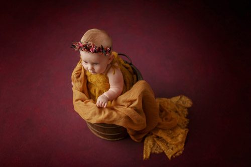 Baby with floral crown sitting in golden fabric on a rustic wooden barrel.