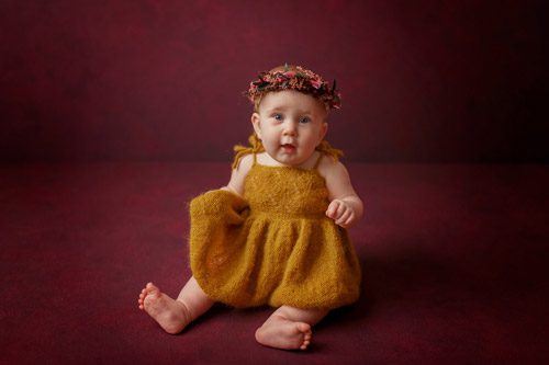 Curious baby sitting upright in a golden dress with a delicate floral headband.