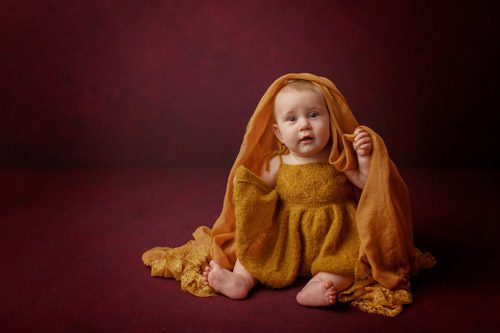 Infant draped in golden fabric, looking thoughtfully while seated.