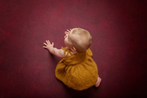 Baby in a playful pose on maroon background, smiling and dressed in golden yellow.