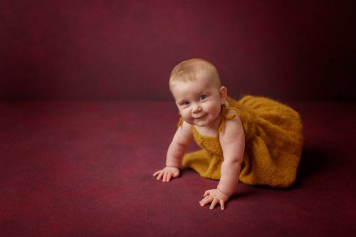 Baby gazing upward, dressed in a golden outfit against a rich maroon backdrop.<br />
