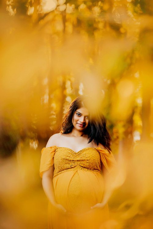 Canberra maternity photographer captures a serene portrait of a pregnant woman in a golden gown, surrounded by the warm hues of autumn foliage