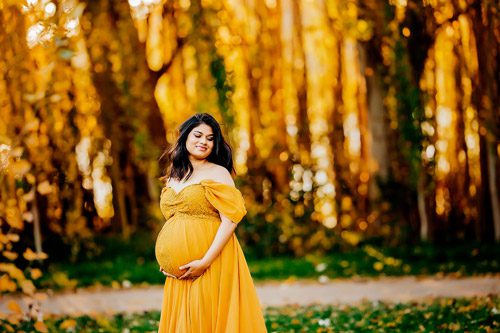 Pregnant woman in a golden maternity gown, standing among golden autumn leaves, captured by a Canberra maternity photographer.